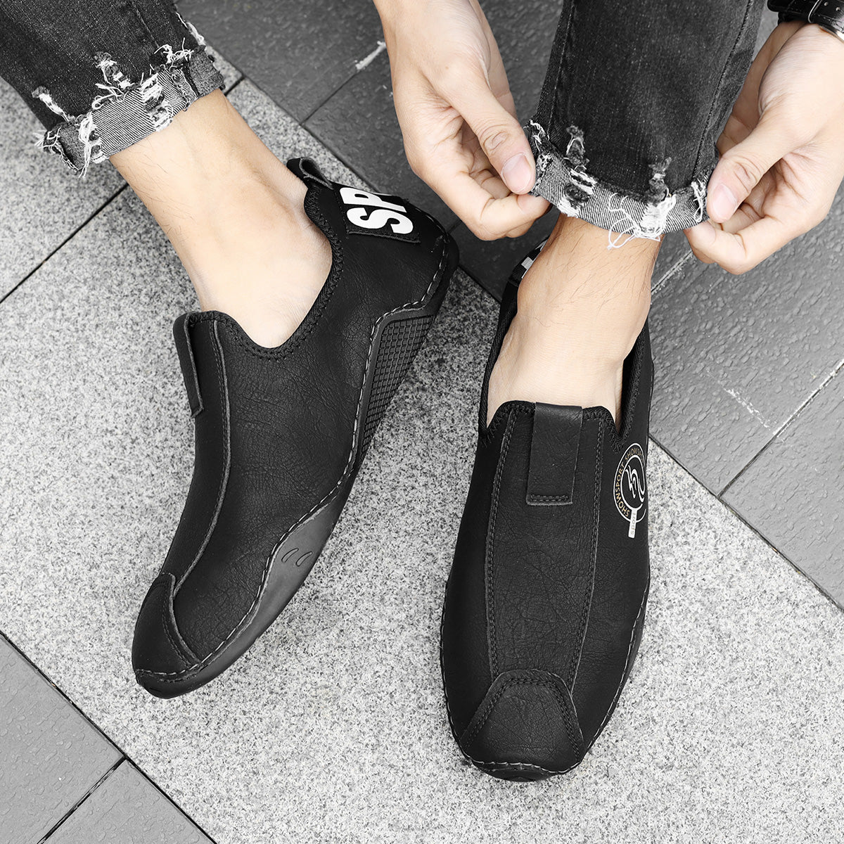 Person wearing black slip-on shoes with a brand logo on a tiled floor.